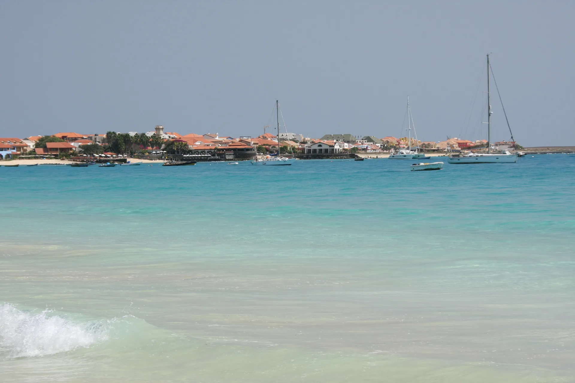 Turquoise shallows of Santa Maria bay on Sal, looking across to the town's low terracotta-roofed buildings and palms with sailing yachts anchored offshore.