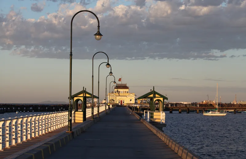 St Kilda Pier at golden hour, its curved green lamp posts and white railings leading along the boardwalk to the yellow kiosk at the end, with a sailing boat moored on Port Phillip Bay