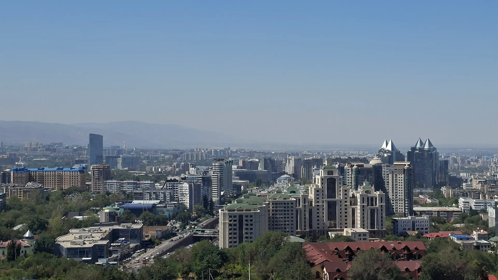 The modern Almaty skyline of mid-rise apartment blocks and glass towers seen from the Kok-Tobe hill, with the foothills of the Tian Shan mountains fading into the haze behind