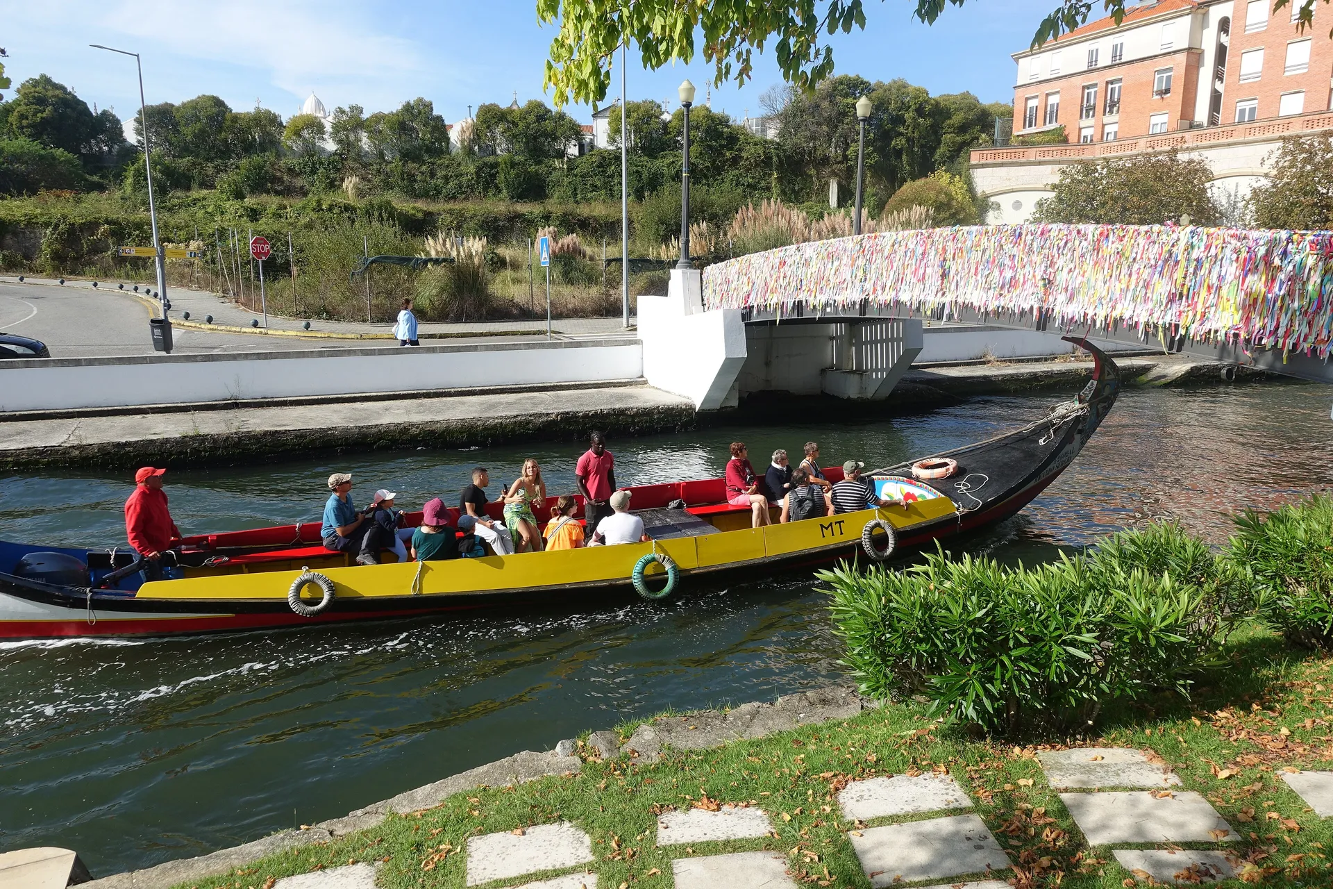 A brightly painted yellow-and-red moliceiro boat carrying visitors along a canal in Aveiro, Portugal, with a ribbon-covered footbridge and Art Nouveau buildings behind