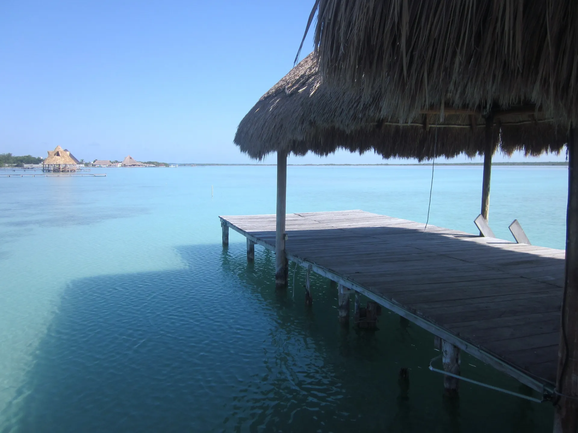 The turquoise water of the Lagoon of Seven Colors at Bacalar, Mexico, with a wooden pier under a thatched palapa roof and palapa huts along the far shore under a clear blue sky