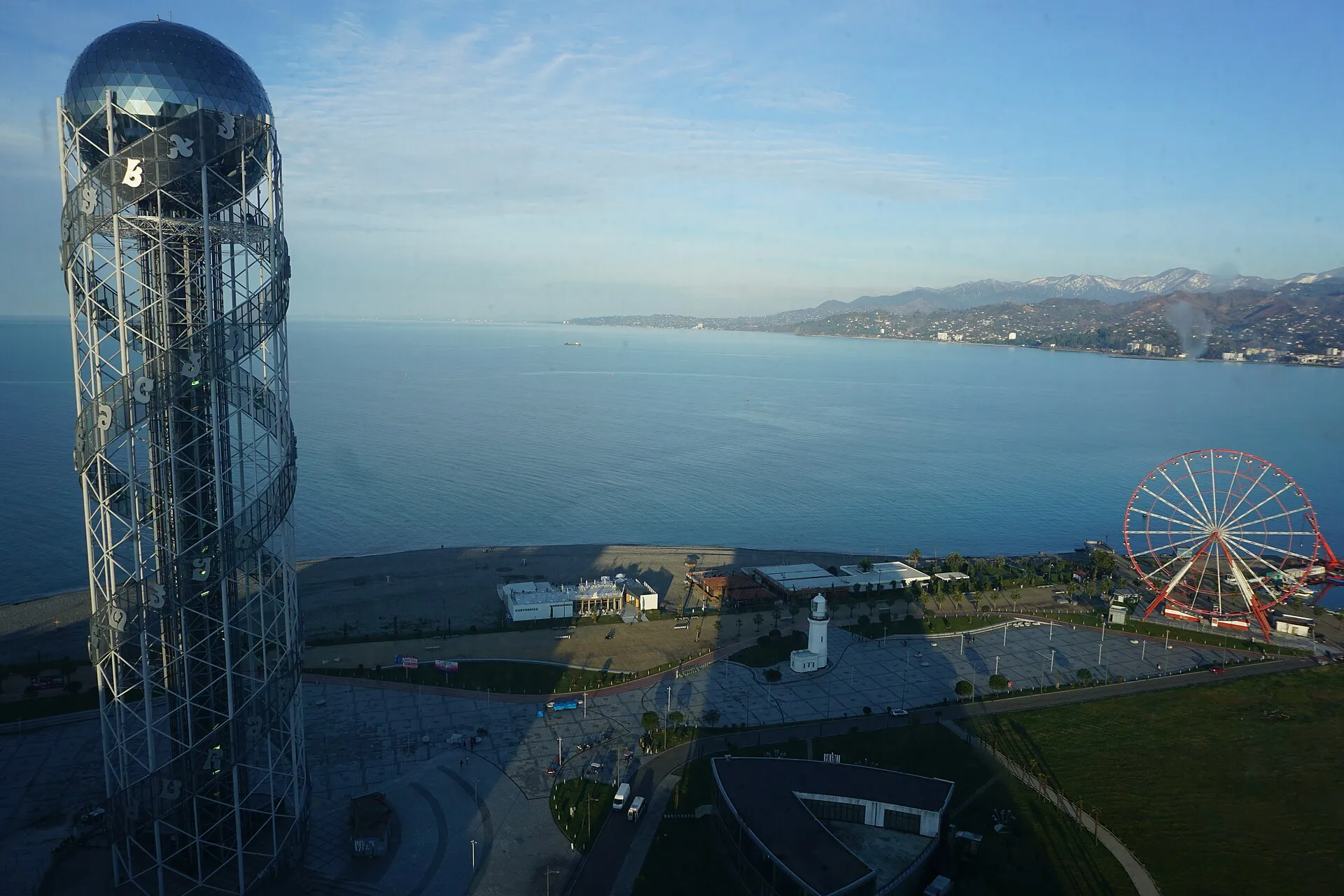 Batumi waterfront viewed from the Alphabetic Tower, showing the Black Sea, Ferris wheel, lighthouse, and snow-capped Caucasus mountains