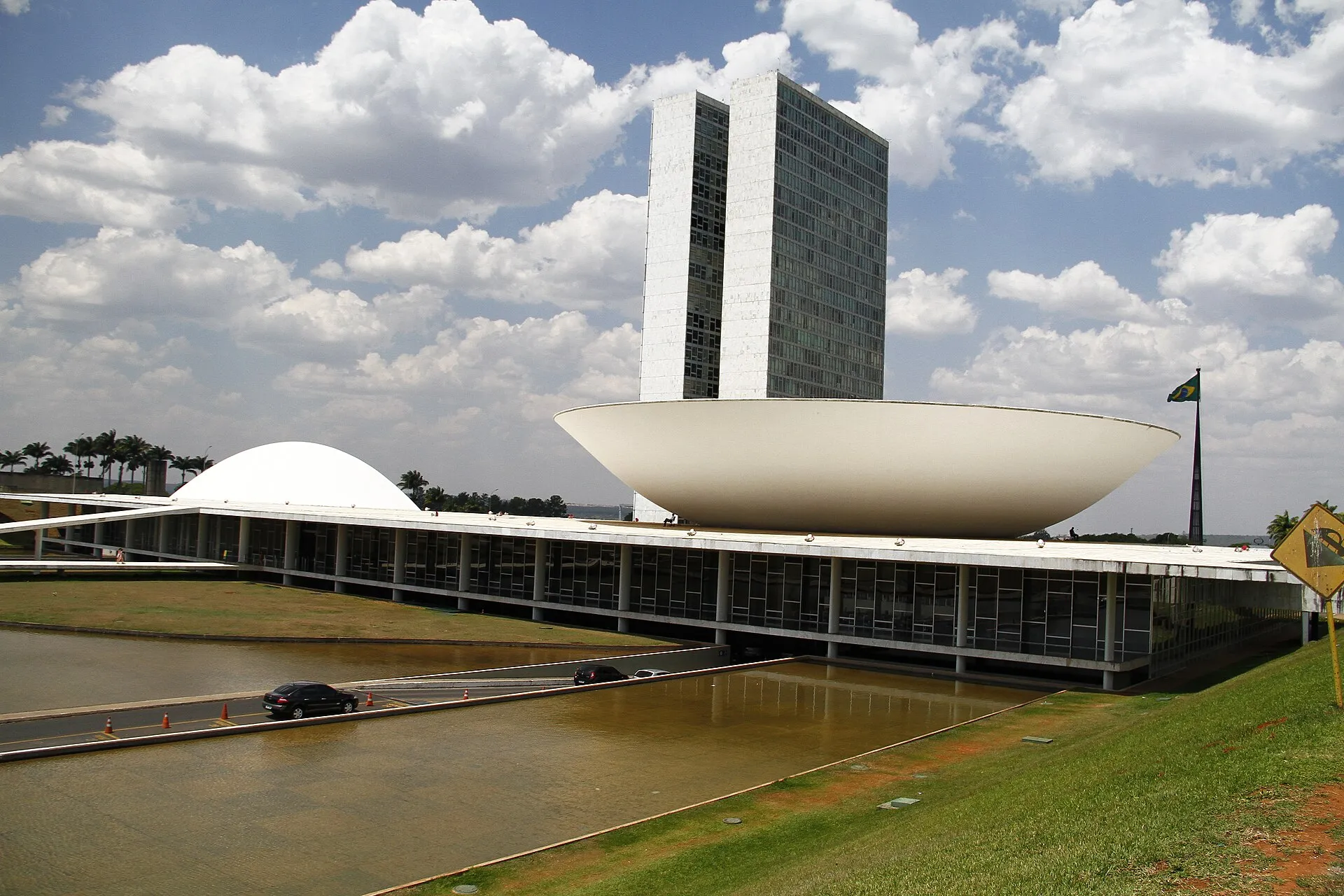 The white bowl and dome of the National Congress designed by Oscar Niemeyer, with its twin towers and reflecting pool under a bright cloudy sky in Brasilia