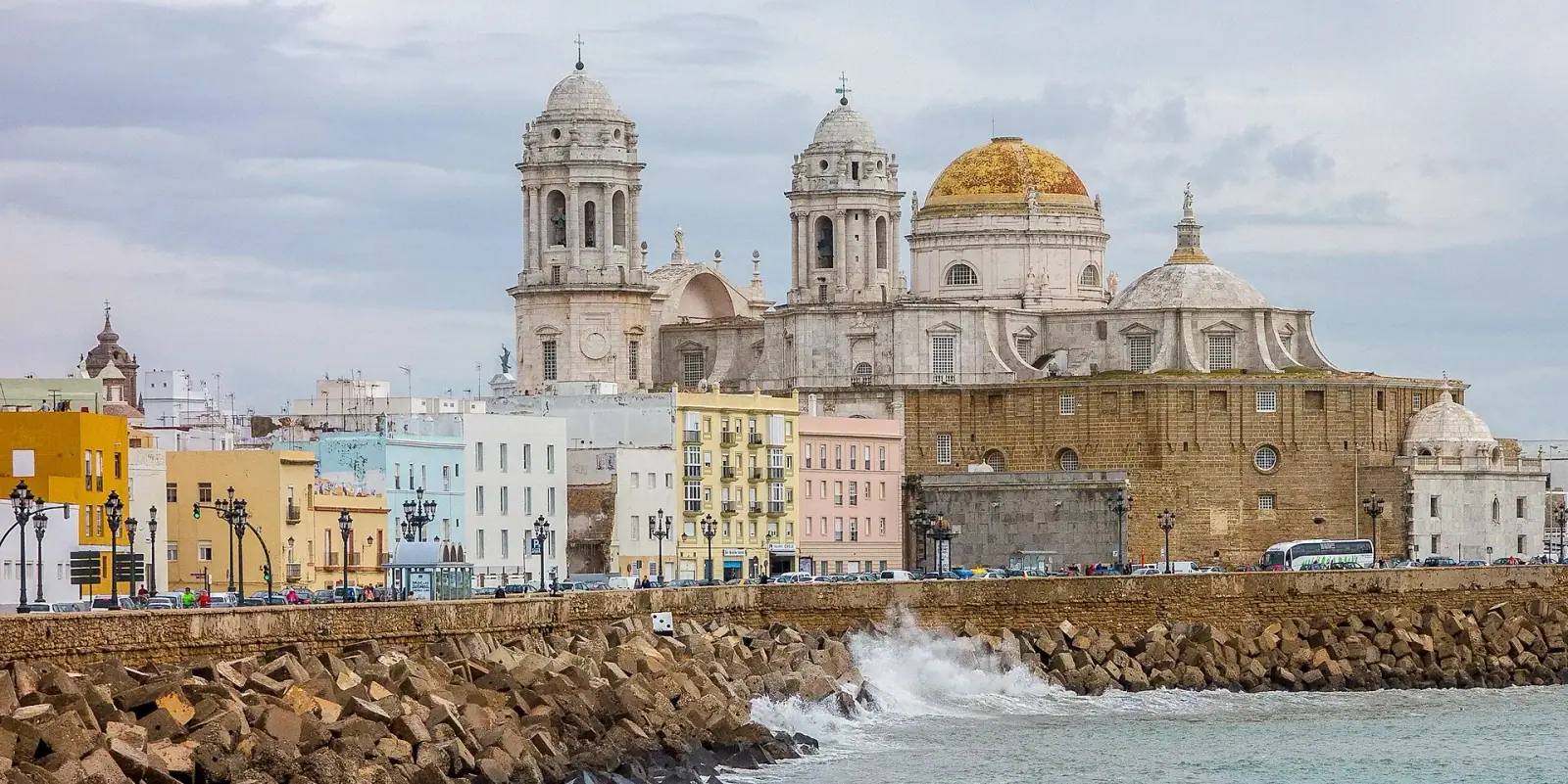 Cádiz Cathedral with golden dome rising above colourful seafront facades, with ocean waves in the foreground
