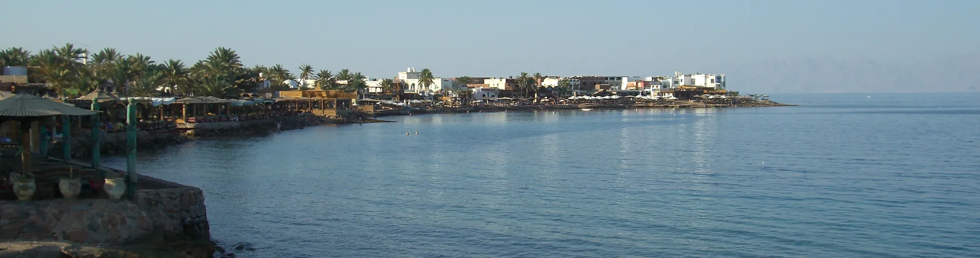 The calm blue water of Masbat Bay in Dahab, the curving Red Sea waterfront lined with palm-shaded beach cafes and low white buildings on the Sinai coast
