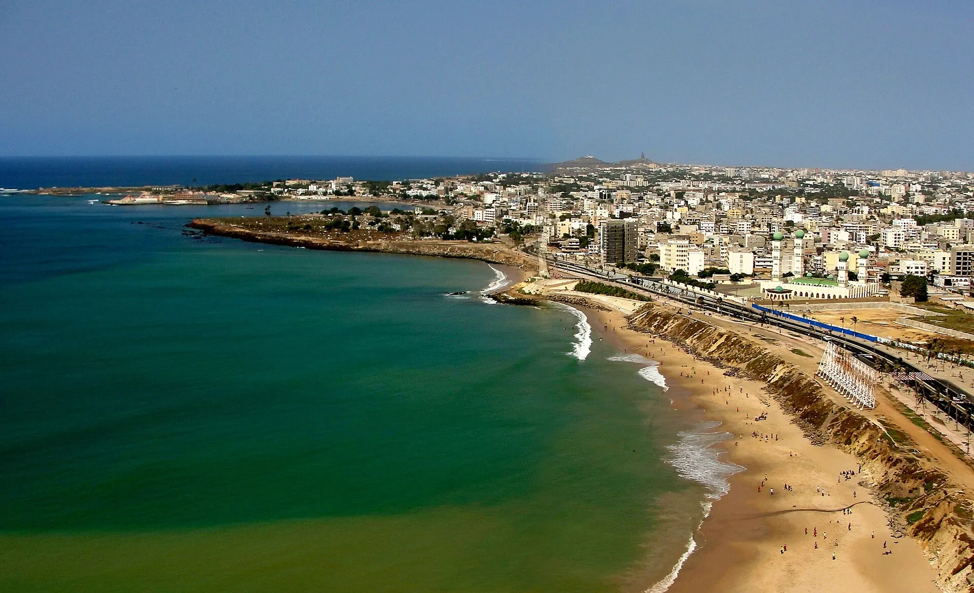 Aerial view of Dakar coastline with teal Atlantic Ocean, sandy beach, city skyline, and the African Renaissance Monument on the horizon