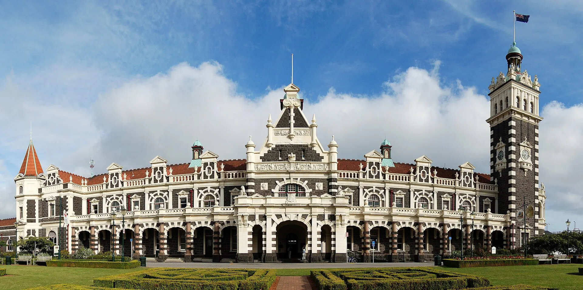 The ornate Edwardian facade and clock tower of Dunedin Railway Station in New Zealand under a blue sky with formal gardens in front.