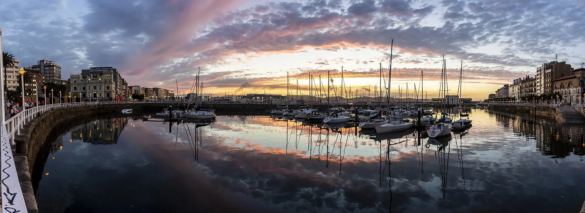 Gijón marina at sunset with sailboats reflected in calm water and city buildings on both sides