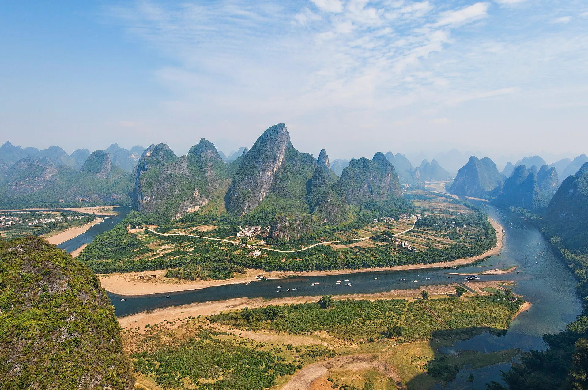 Limestone karst peaks crowd the banks of the Li River south of Guilin, with farmland, a riverside village and tour boats below a hazy blue sky