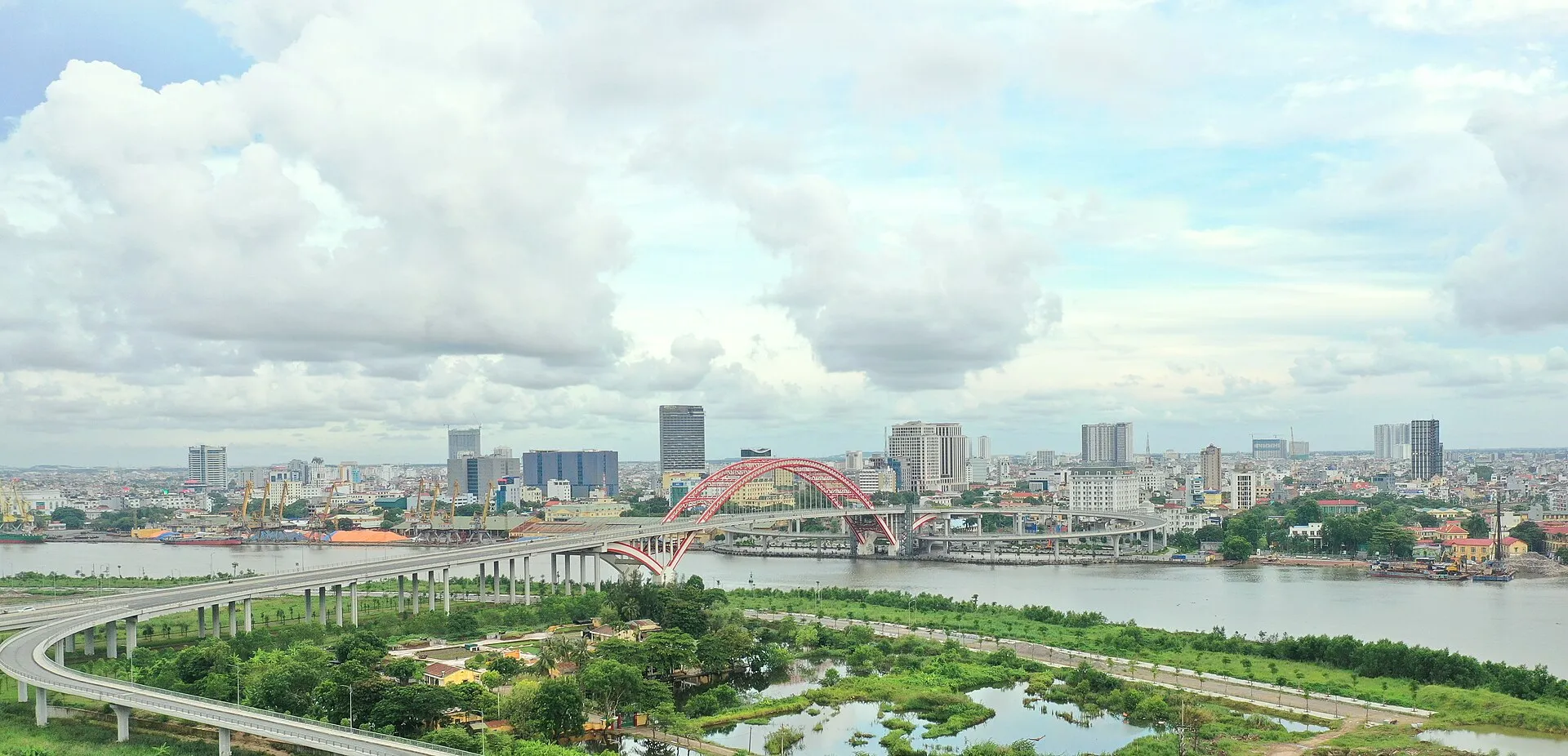 The Hai Phong skyline in Vietnam seen across the Cam River, with the red arch of Hoang Van Thu Bridge and port cranes below tall white clouds.