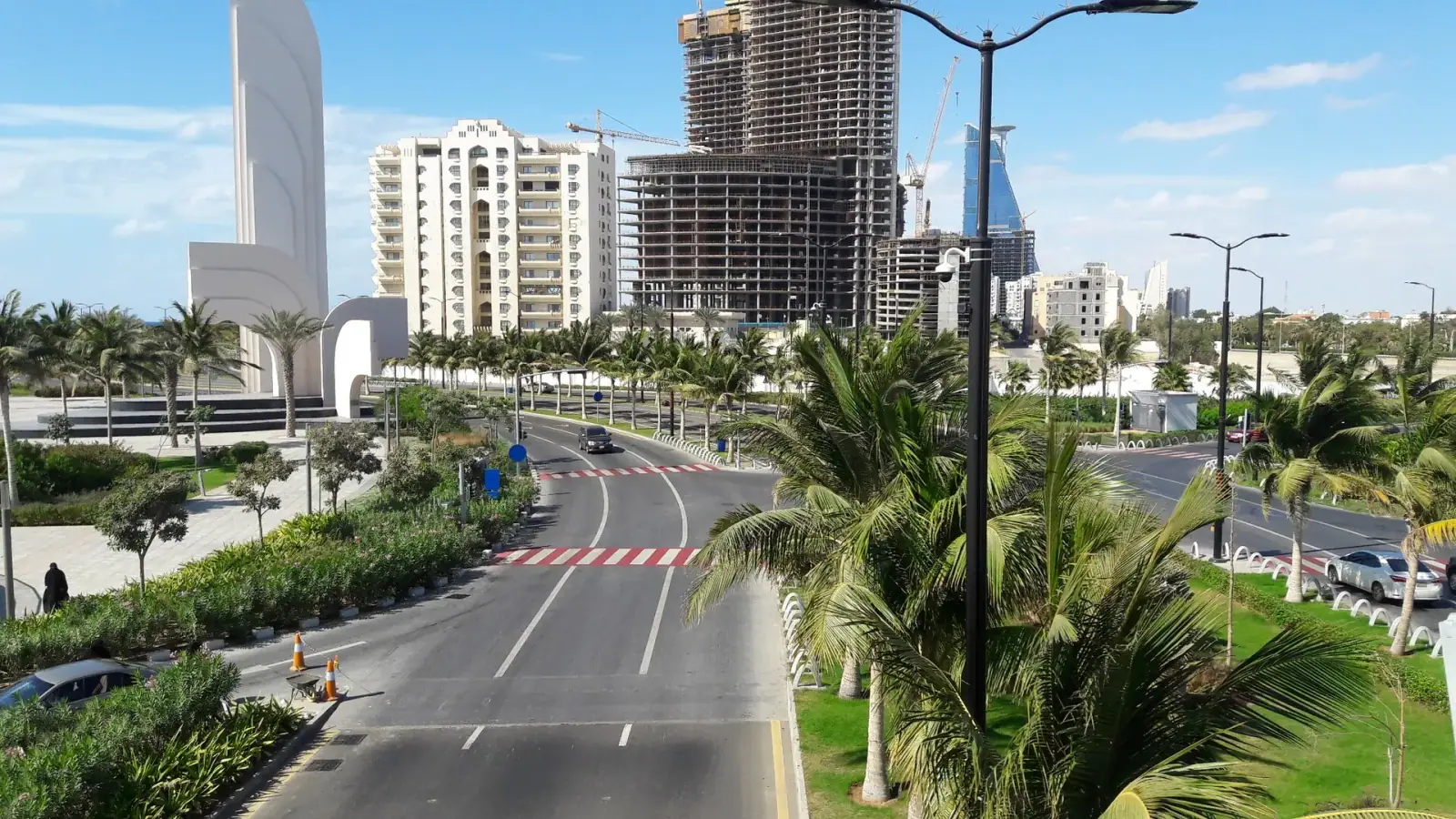 Jeddah Corniche boulevard with palm trees, white arch monument, and modern towers under a blue sky
