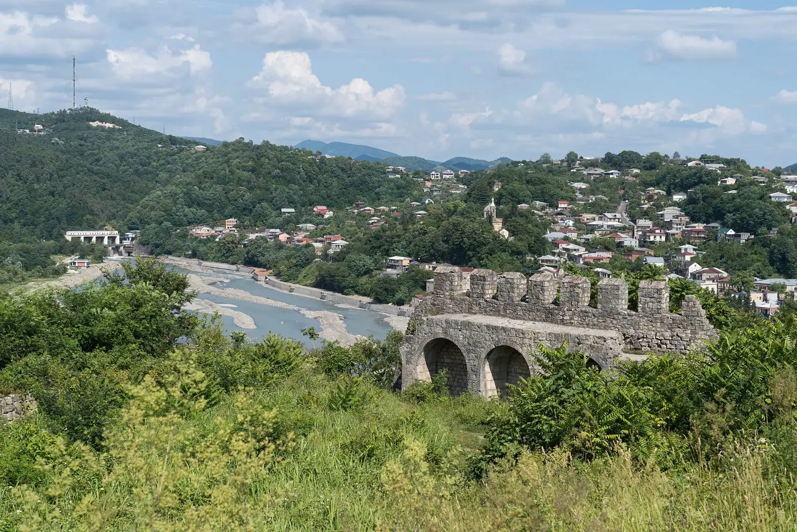 View of Kutaisi from the Ukimerioni Fortress, showing the ancient stone bridge ruins over the Rioni River and the city spreading across the green hills