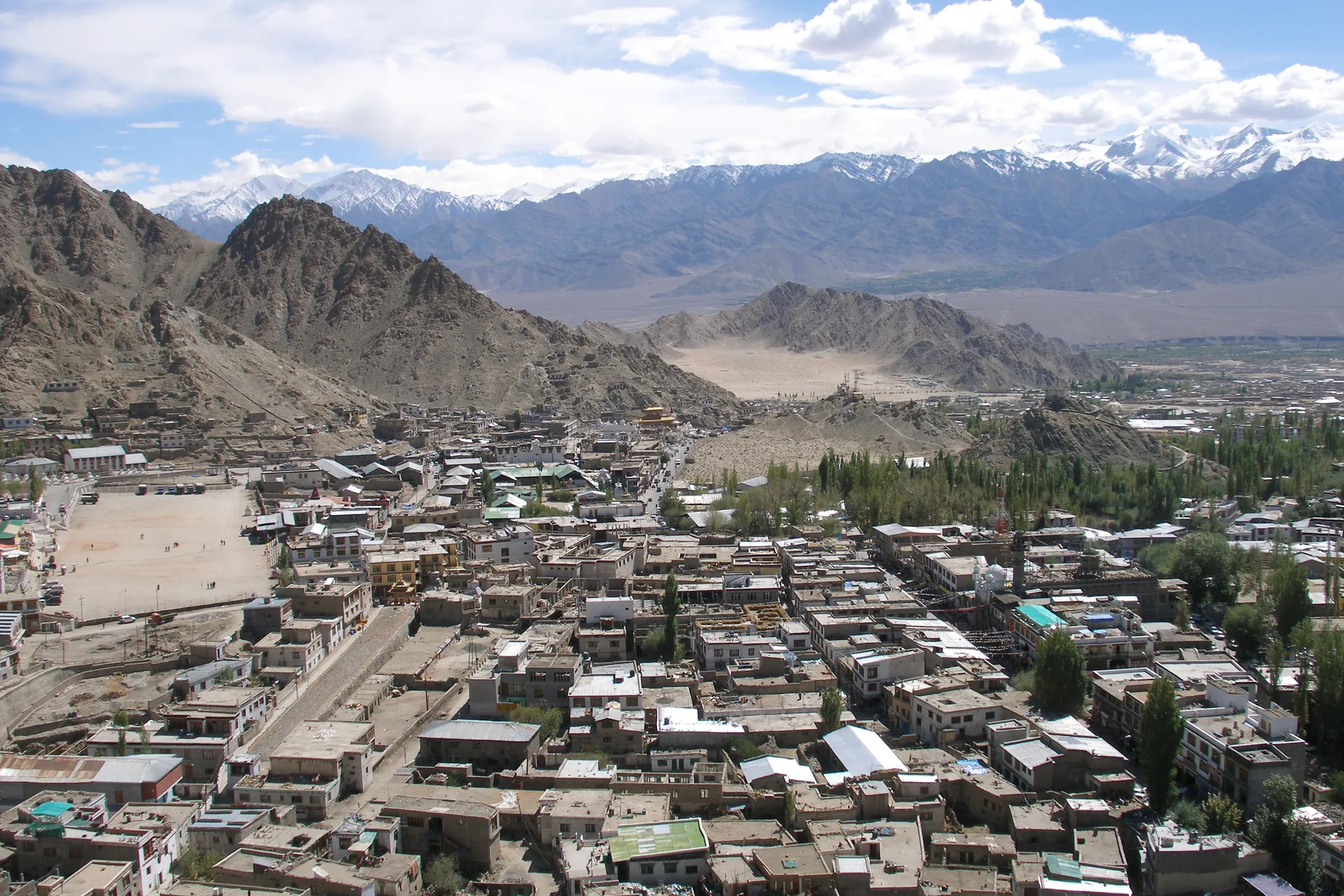 The high-altitude town of Leh spreading across a desert valley in Ladakh, ringed by bare brown mountains with the snow-capped peaks of the Himalayas rising behind under a blue sky