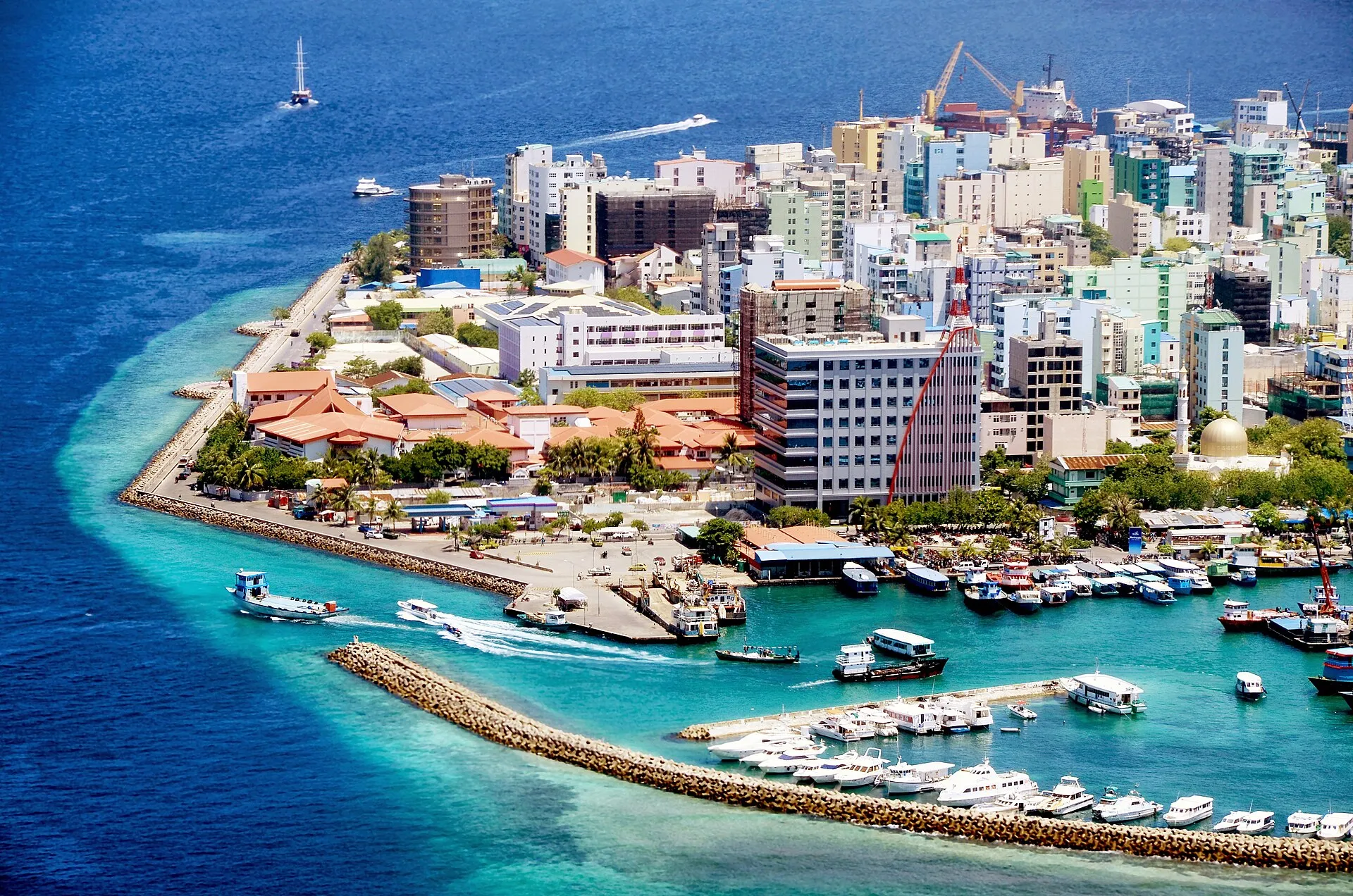 Aerial view of the dense island city of Male ringed by turquoise lagoon water, harbour breakwaters and moored boats.