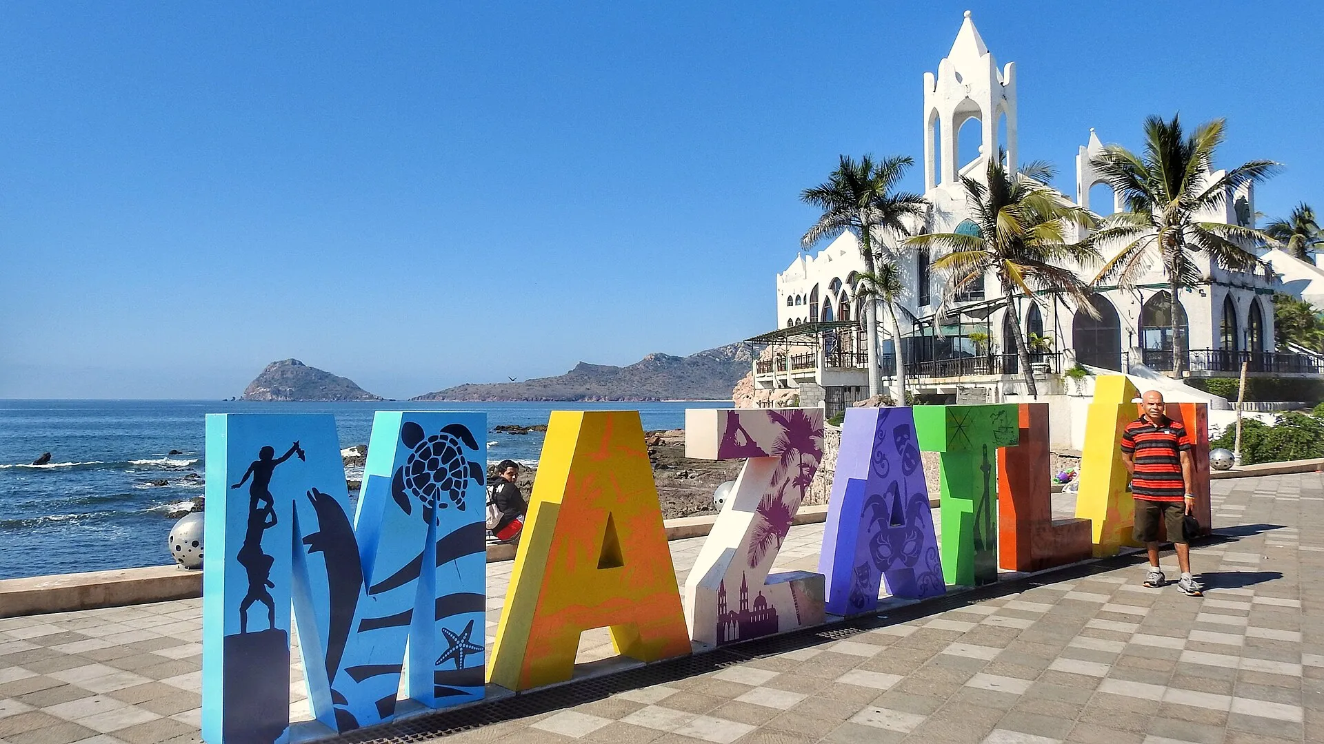 Colorful MAZATLAN sign on the malecón with Valentino's building, palm trees, and the Pacific Ocean