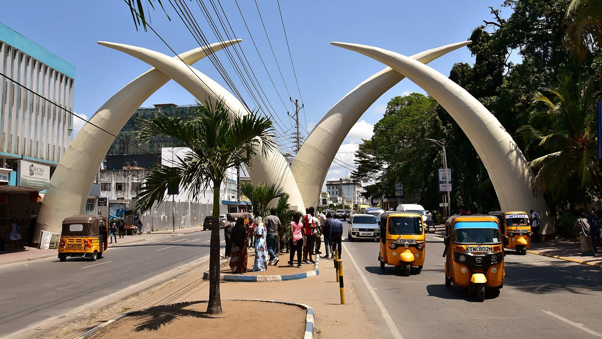 The giant white Mombasa Tusks arching over Moi Avenue with yellow tuk tuks passing beneath on a sunny day