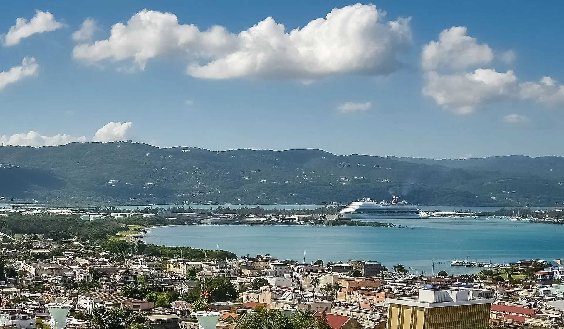 Elevated view over the rooftops of Montego Bay toward the turquoise harbour, a docked cruise ship and the green hills of Jamaica beyond.