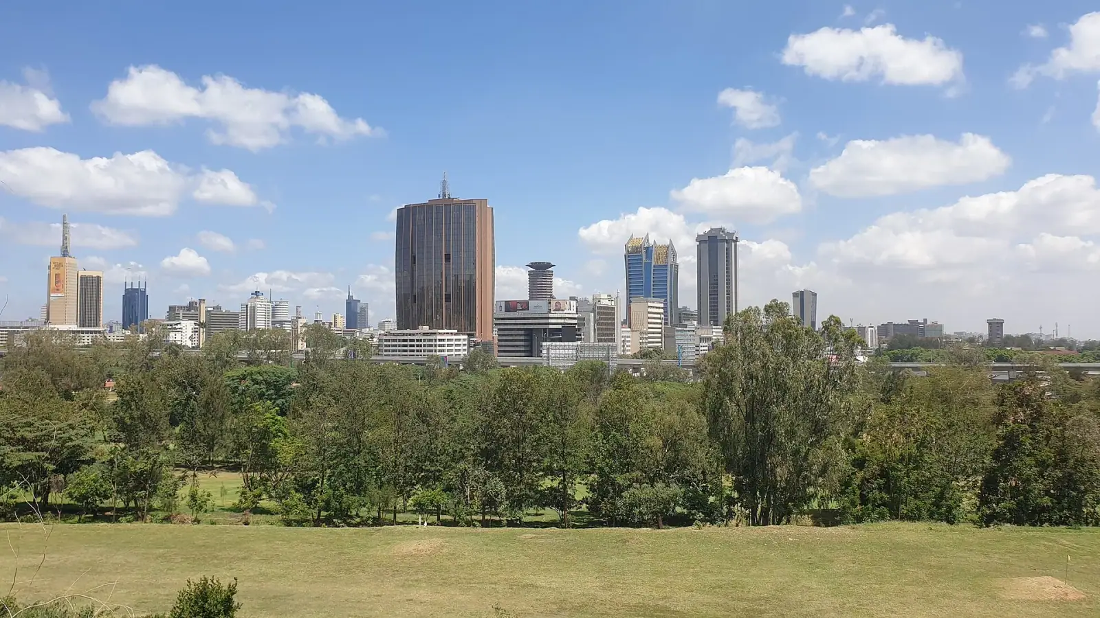 Nairobi CBD skyline viewed from Uhuru Park, with landmark skyscrapers rising above green trees under a blue sky