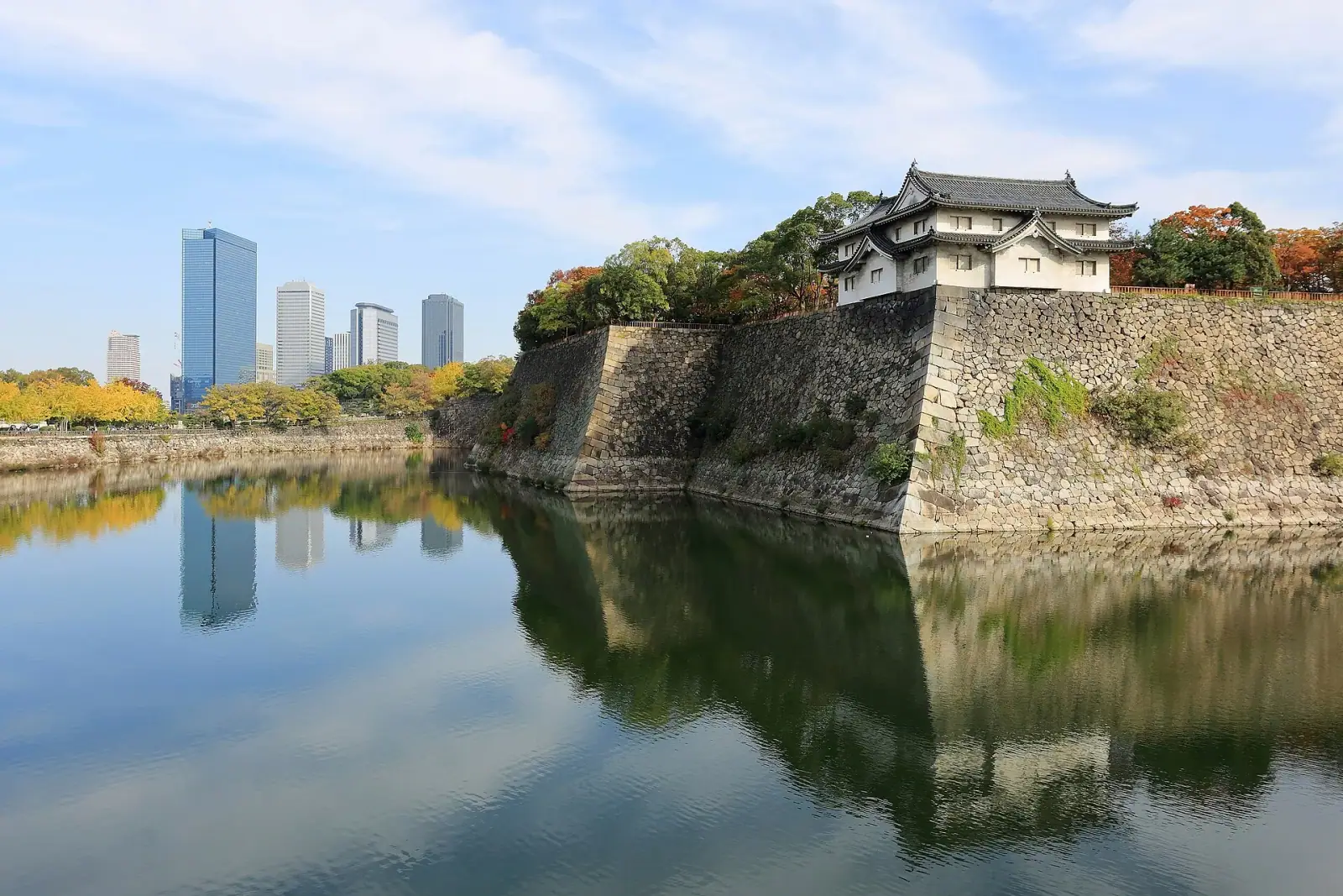 Osaka Castle outer moat with stone walls and turret reflected in water, modern Business Park skyscrapers in the background