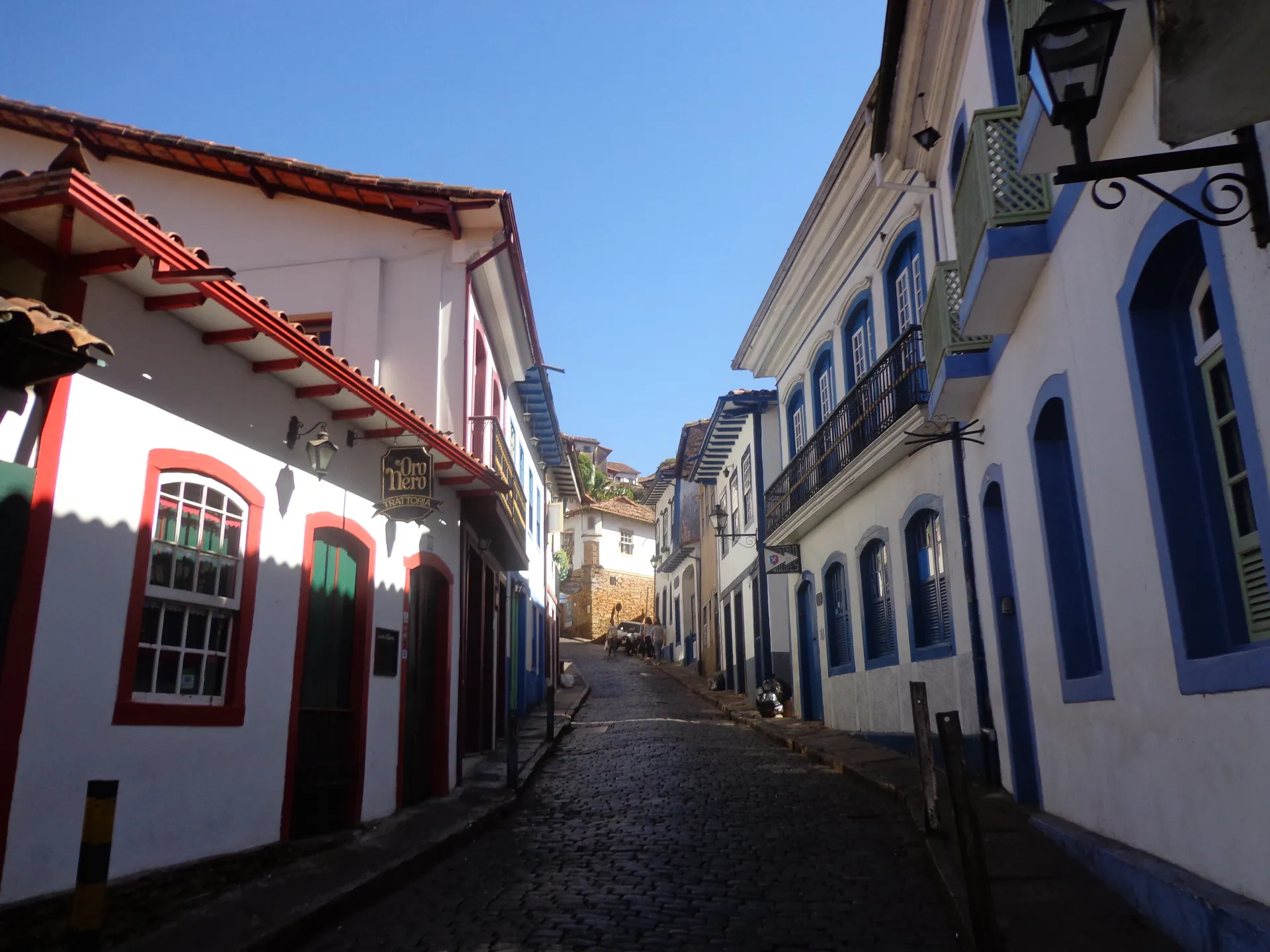 A steep cobbled colonial street in Ouro Preto lined with white baroque houses trimmed in red, blue and green, a wrought-iron street lamp overhead under a clear blue sky