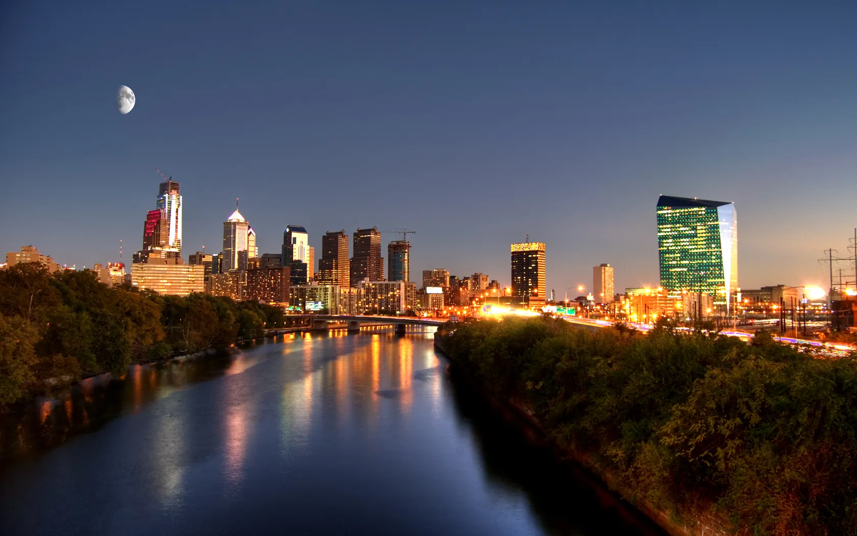 The Philadelphia skyline at dusk reflected in the Schuylkill River, with a moon rising over the lit downtown towers