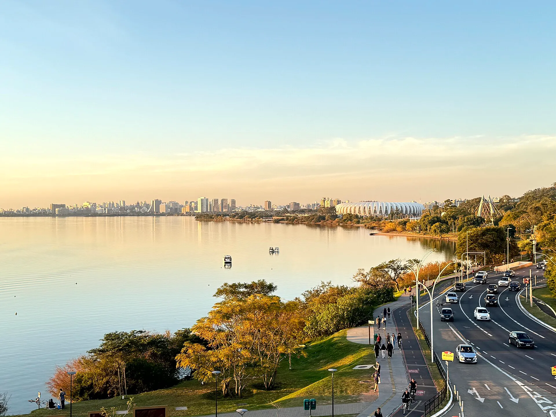 Sunset over the Guaiba waterfront in Porto Alegre, with the city skyline and the Beira-Rio stadium across the calm water and cyclists on the riverside promenade