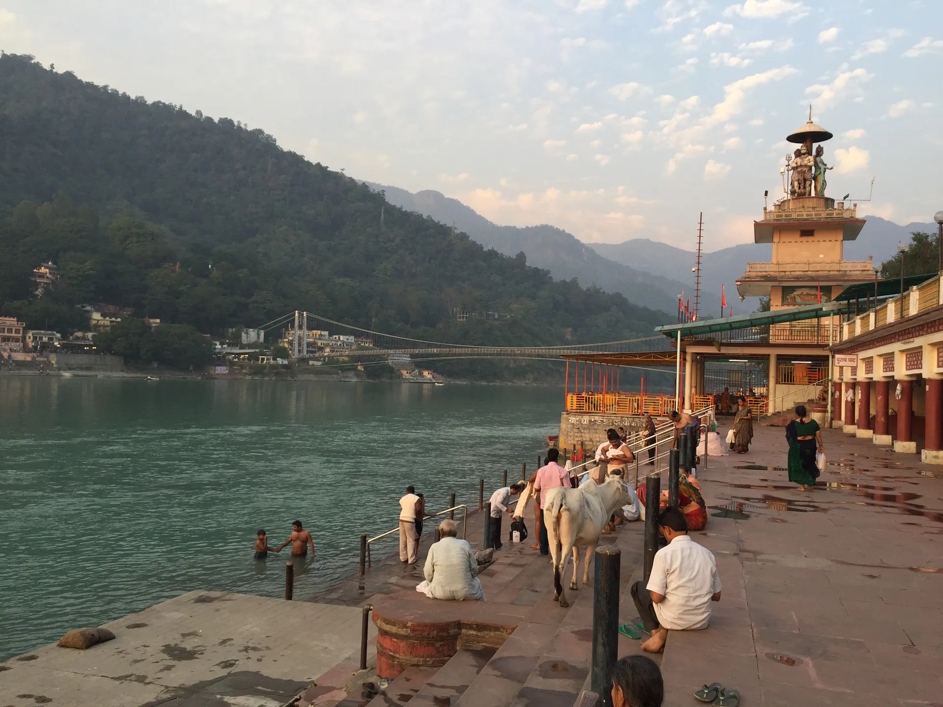 Pilgrims bathing at a stone ghat on the Ganges in Rishikesh at golden hour, a riverside temple topped by a Shiva statue on the right and a suspension footbridge crossing to the forested Himalayan foothills behind