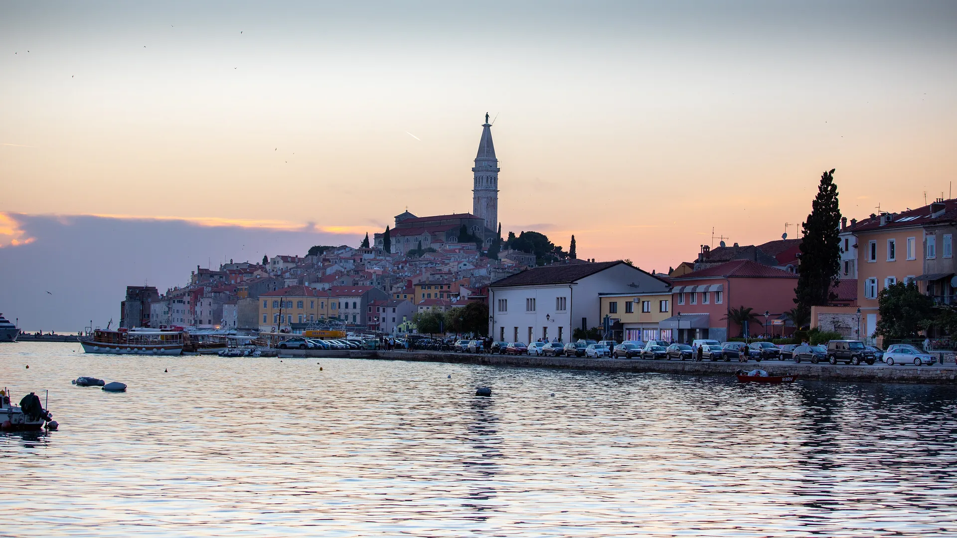 The old town of Rovinj on its peninsula at sunset, pastel houses rising to the tall Venetian bell tower of St Euphemia, seen across the calm Adriatic with boats along the waterfront