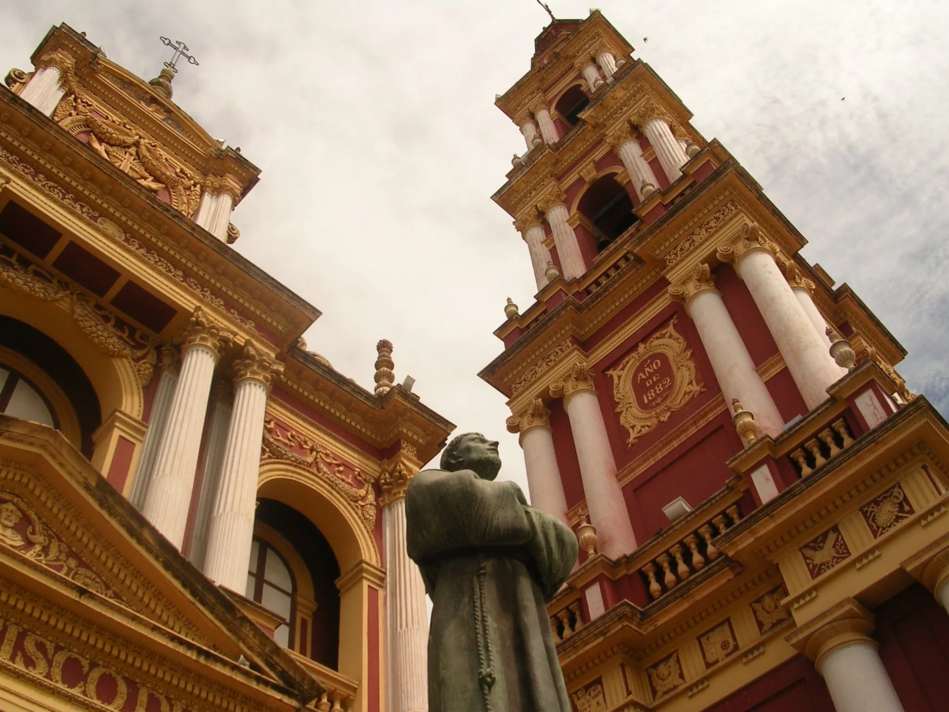 The ornate terracotta-red and ochre-gold facade and bell tower of the San Francisco church in Salta, Argentina, seen from below with a friar statue in front