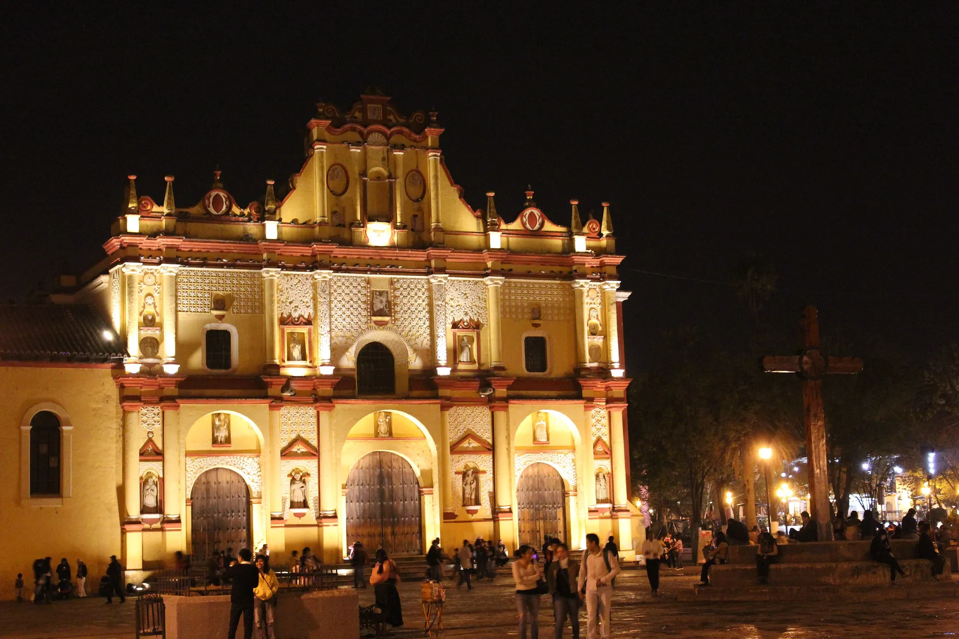 The ornate yellow-and-red baroque facade of the cathedral of San Cristobal de las Casas floodlit at night, with people gathered in the plaza in front
