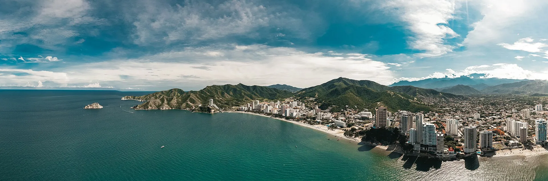 Aerial panorama of El Rodadero bay, Santa Marta, Colombia, with turquoise Caribbean Sea, crescent beach, white high-rise hotels, and Sierra Nevada mountains in the background