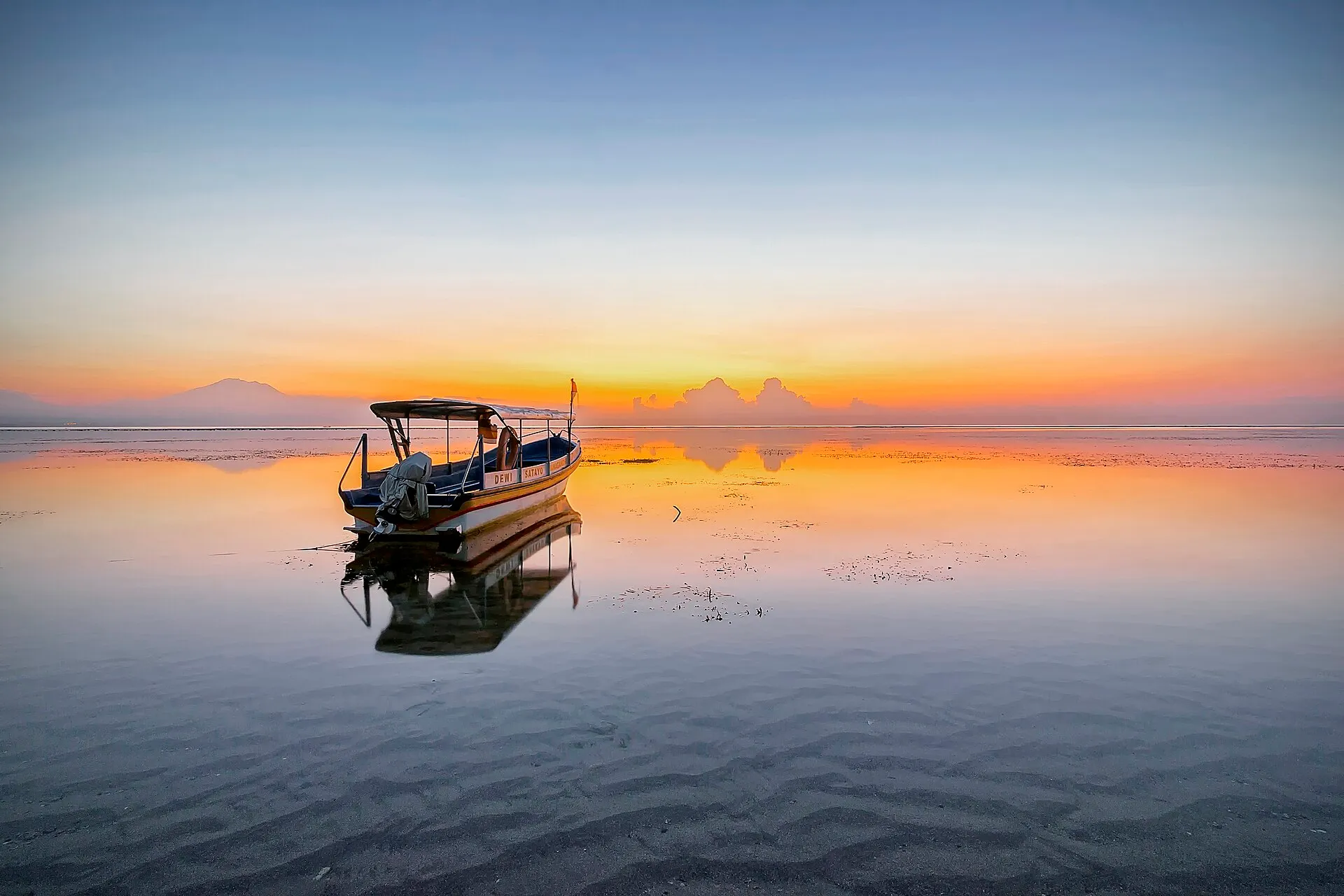 Sunrise over Sanur Beach, Bali, with a traditional boat reflected in calm shallow water and Mount Agung on the horizon