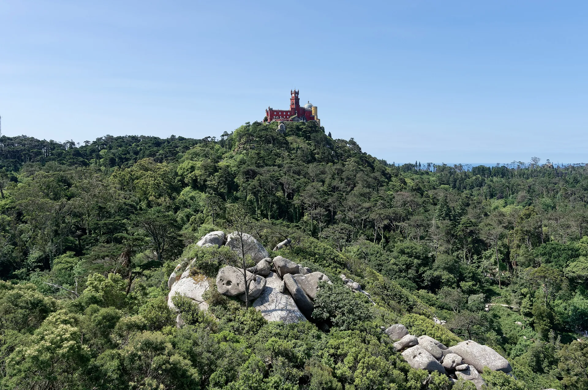 The colourful red-and-yellow romantic Pena Palace perched on a forested hilltop above the green wooded hills of Sintra under a clear blue sky