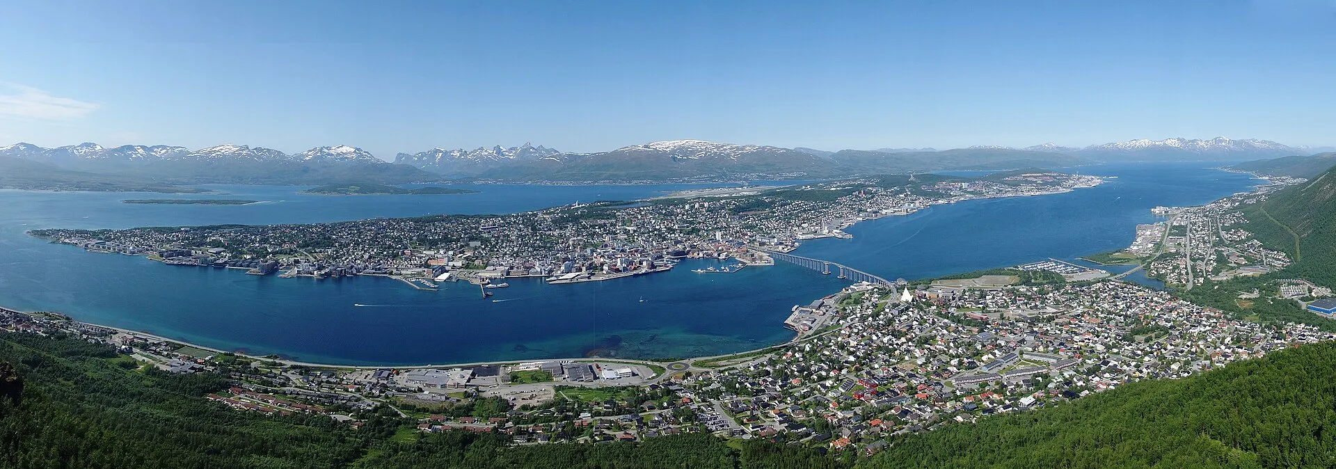 Panoramic view of Tromsø from Fjellheisen, showing the city island, Tromsø bridge, Arctic Cathedral, harbor, and snow-capped mountains under a clear blue sky
