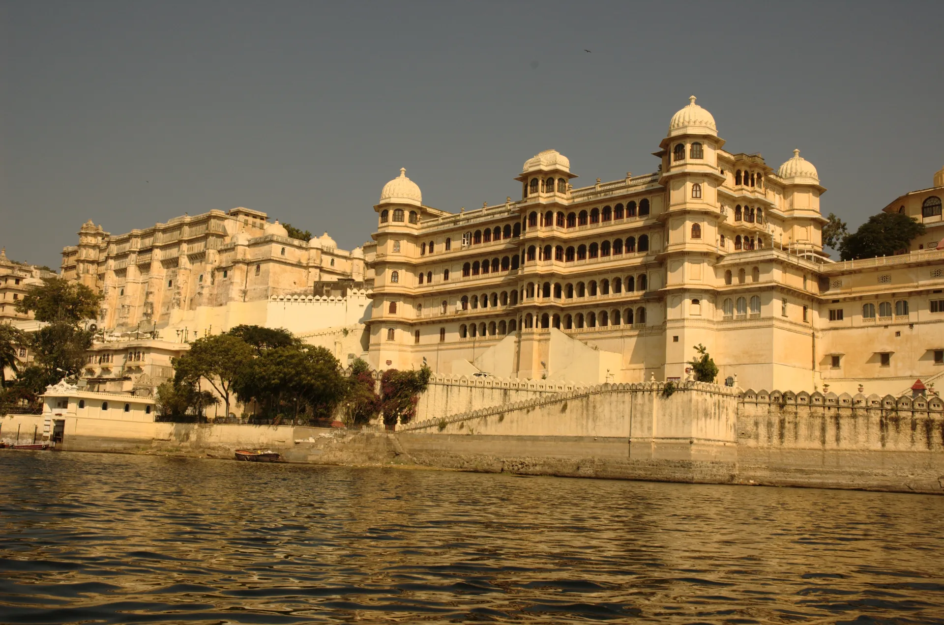 The grand golden City Palace of Udaipur rising above the waters of Lake Pichola, its domed cupolas and tiered facade seen from a boat on the lake