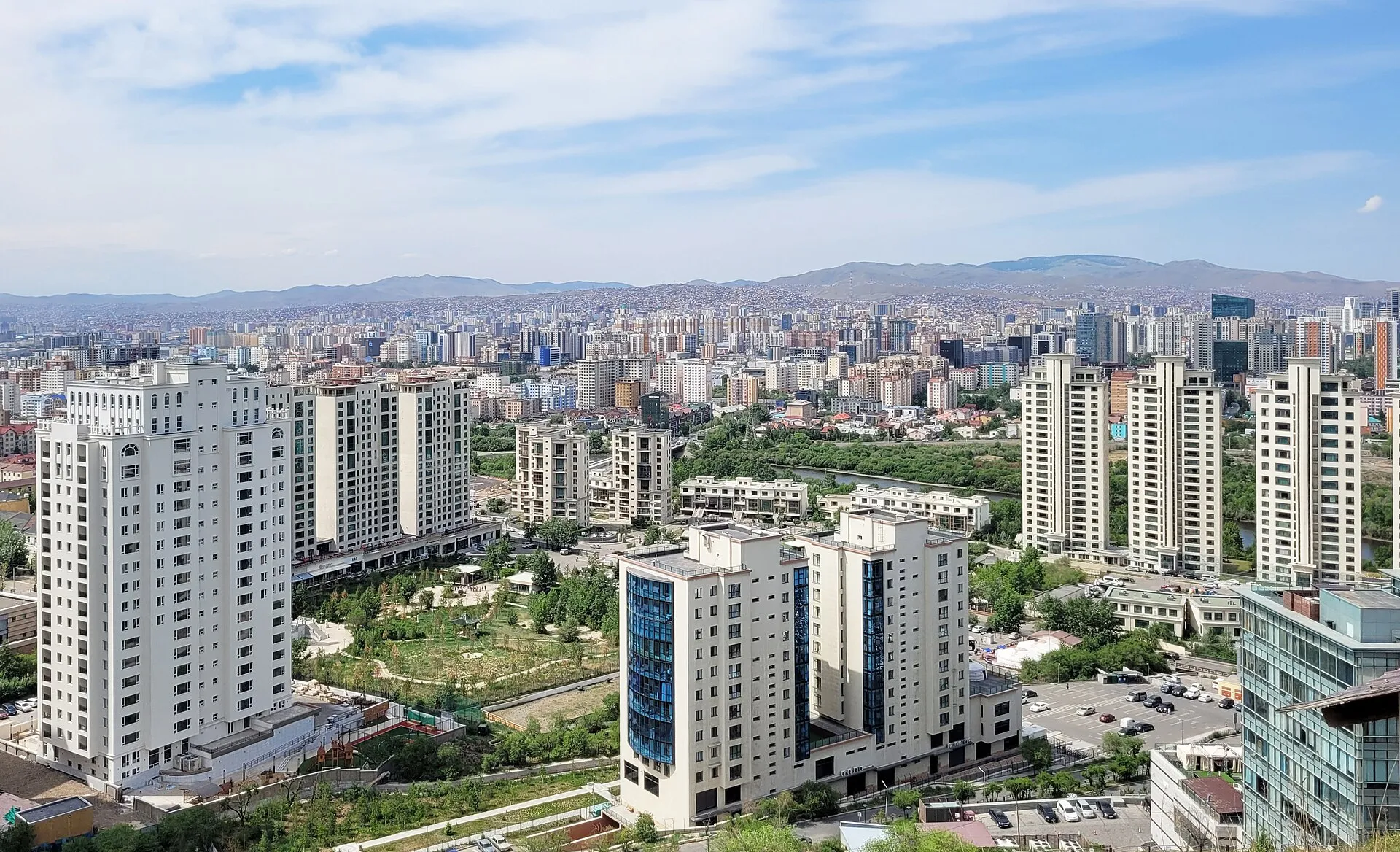 Modern apartment towers and the wide city centre of Ulaanbaatar spreading toward the surrounding Mongolian hills under a blue summer sky.