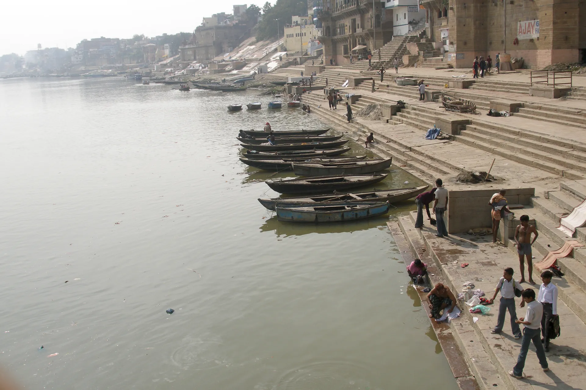 The stone ghats of Varanasi descending to the Ganges at hazy morning light, wooden rowing boats moored along the steps with people bathing and gathering by the river