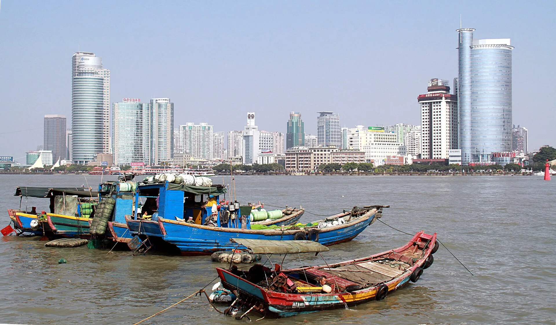 Brightly painted wooden fishing boats moored in the strait in front of the Xiamen waterfront skyline on a clear day, seen from Gulangyu Island