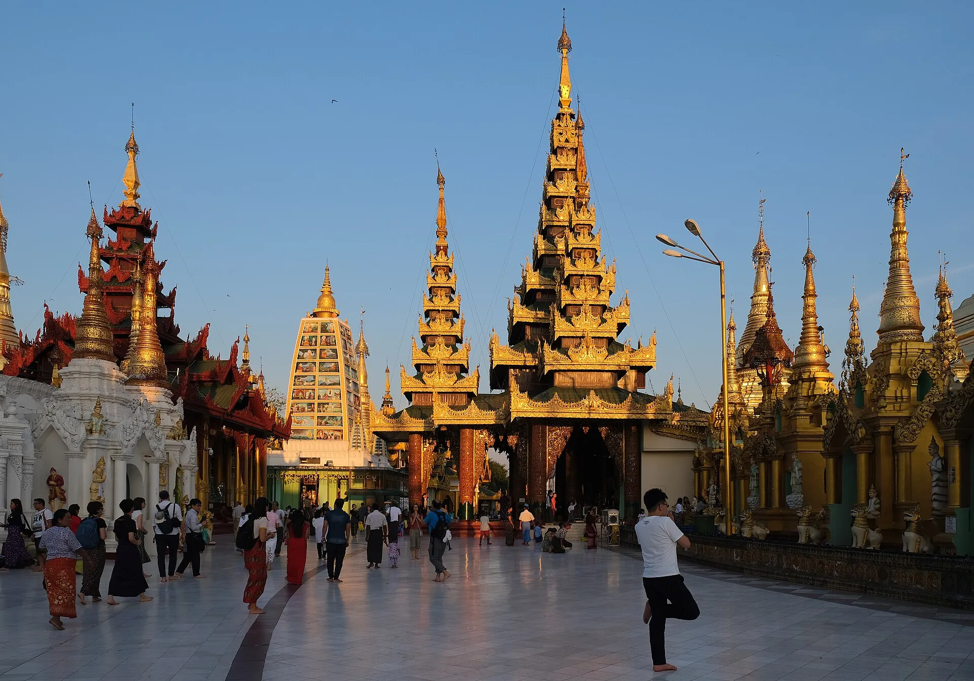 Golden spires and shrines of the Shwedagon Pagoda terrace glowing at golden hour as visitors walk barefoot across the marble plaza in Yangon, Myanmar.