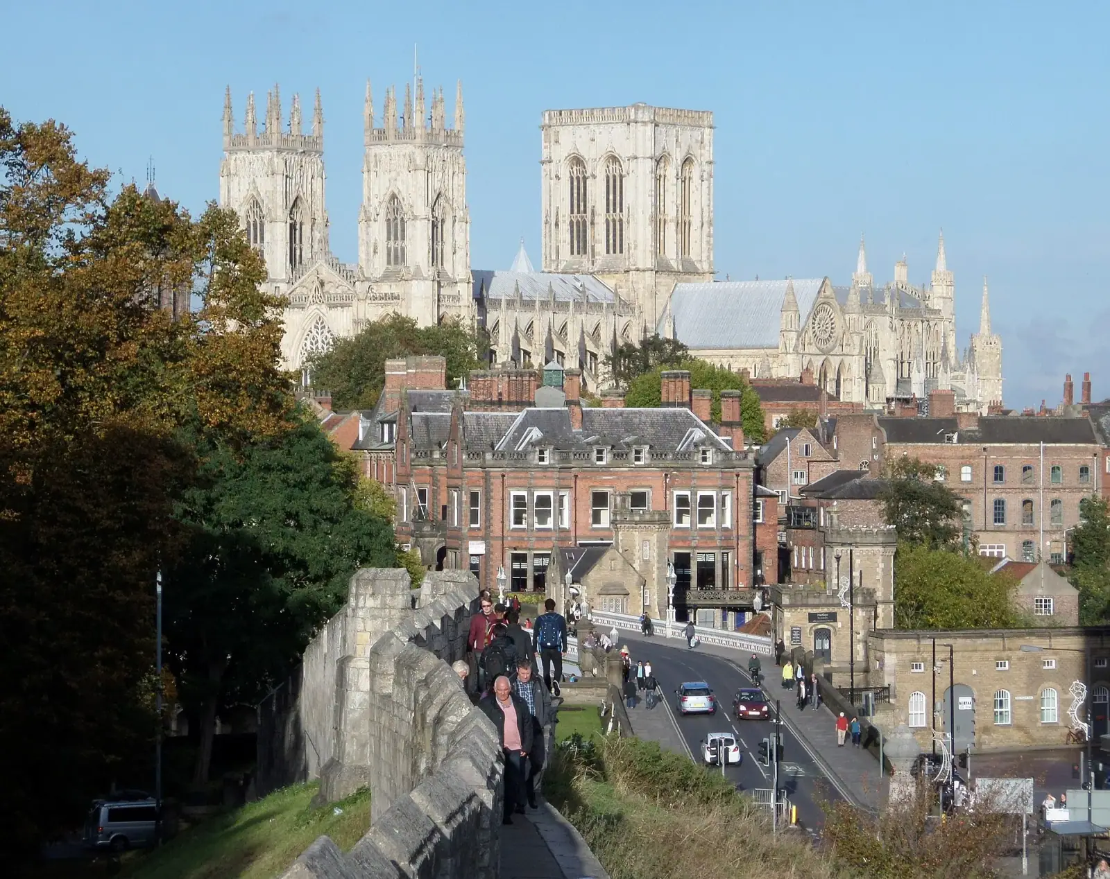 York Minster's twin towers rising above the city skyline, with people walking the ancient city walls in the foreground on a sunny autumn day